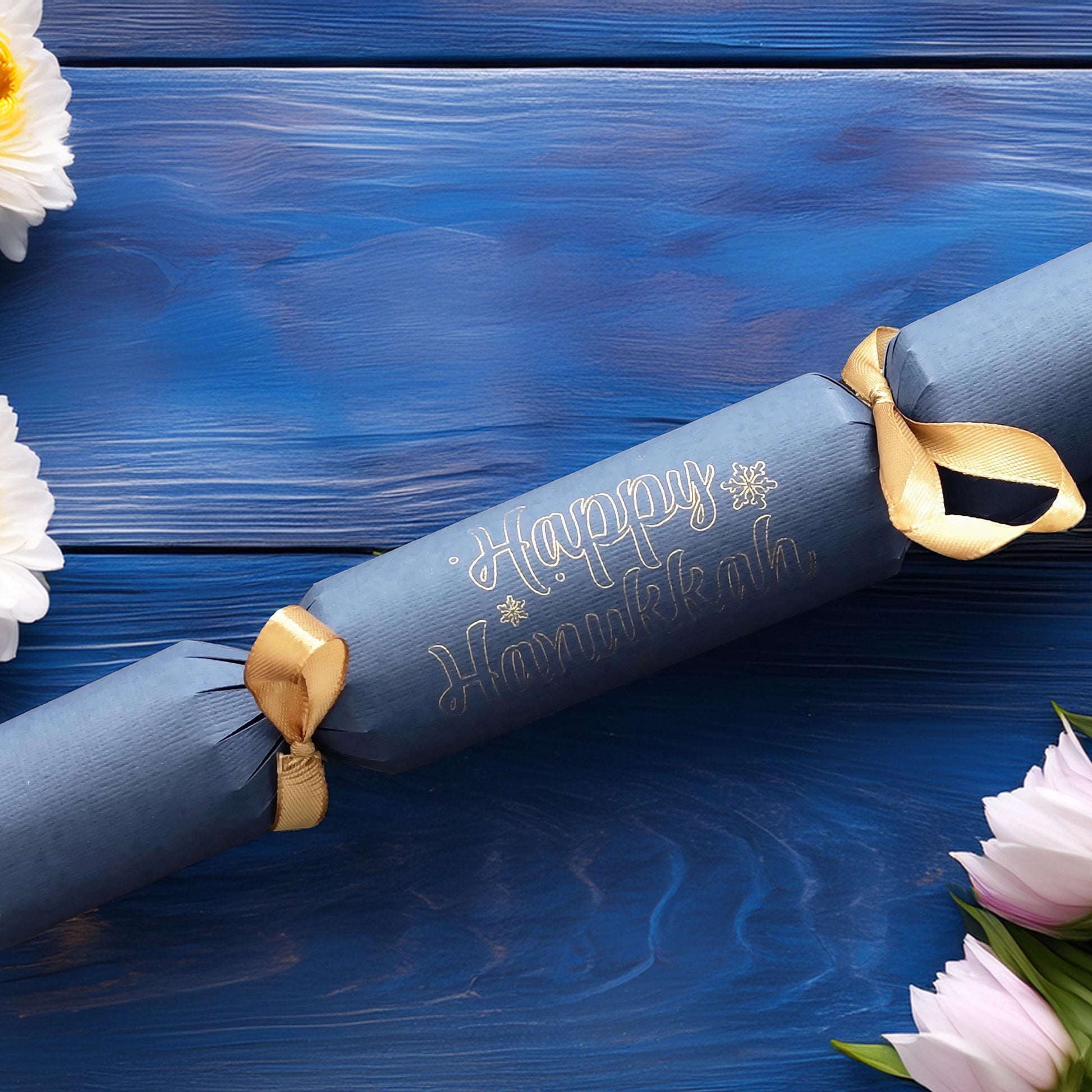 Handmade navy cracker with “Happy Hanukkah” hand-lettered in gold, finished with gold ribbon, styled with white and pink flowers on a blue wood background.