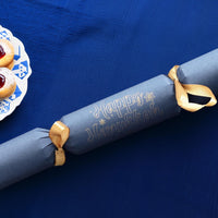 Navy blue cracker with “Happy Hanukkah” in silver pen, tied with gold ribbon, styled on a royal blue tablecloth beside jam-filled pastries.