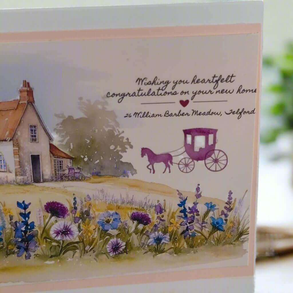 Close-up angled view of a handmade New Home card featuring a countryside cottage, colourful wildflowers, and a purple horse-drawn carriage, with personalised text for a thoughtful housewarming message.
