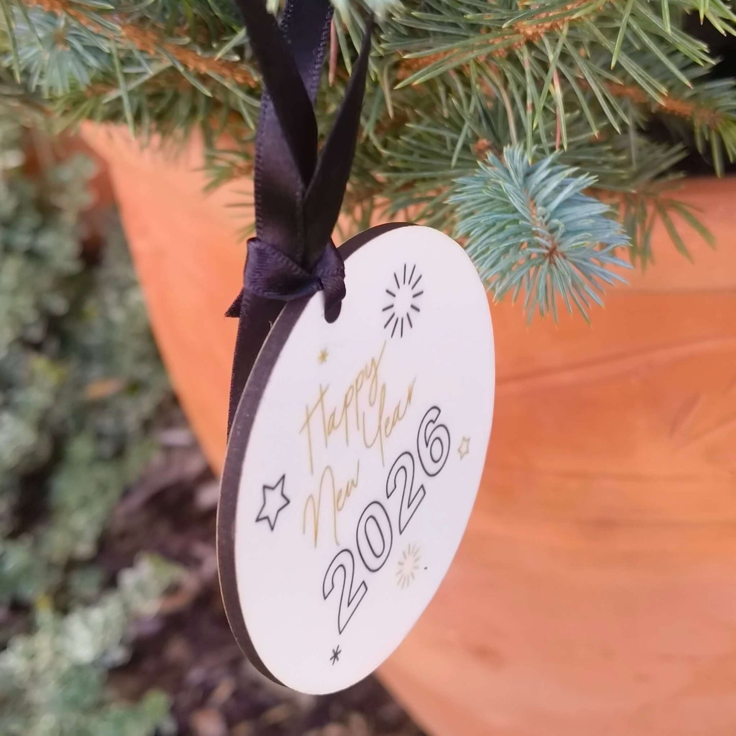 Close-up of a white ceramic New Year bauble with black ribbon and “Happy New Year 2026” design, hanging from a pine branch in a terracotta pot.