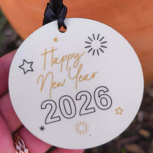 Close-up of “Happy New Year 2026” ceramic bauble with black ribbon, gold and black festive text, photographed in hand with garden background.