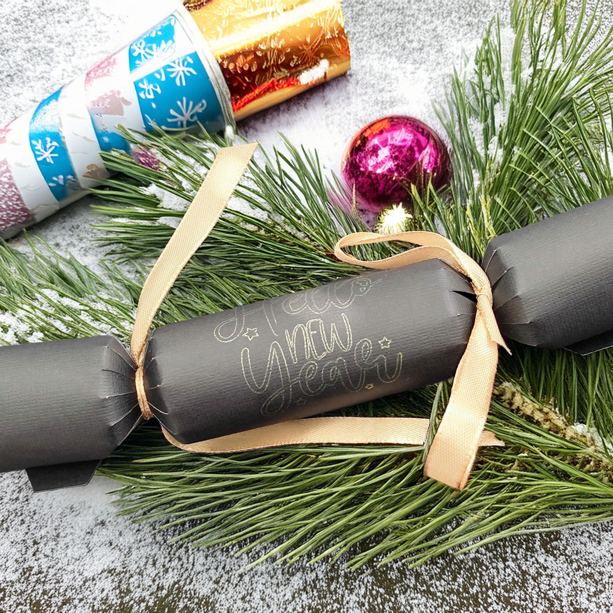 Handmade black New Year cracker tied with gold ribbon, featuring elegant “Happy New Year” lettering. Styled with greenery, baubles, and snow for a festive display.
