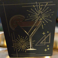 Foil cocktail glass New Year card, front angle, placed on a glittery gold surface with fairy lights and festive bokeh background