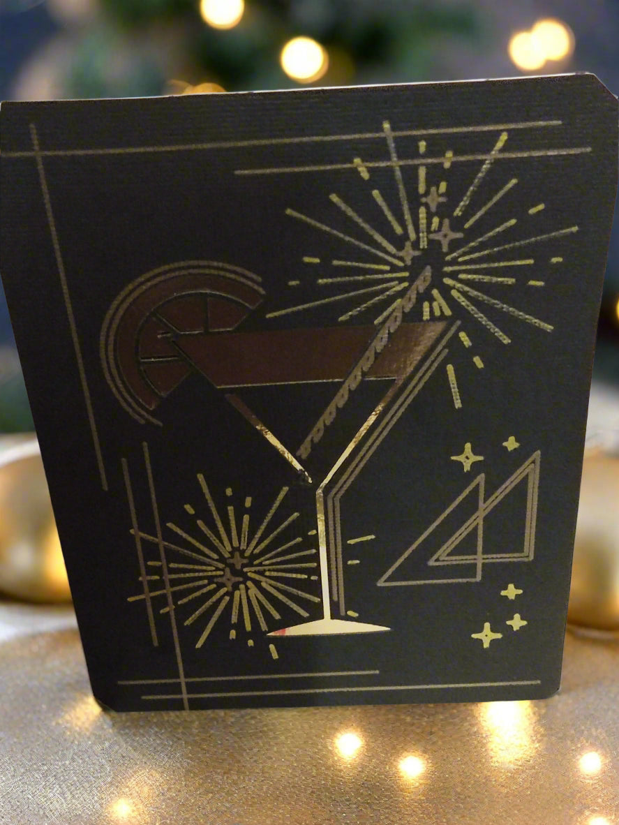Foil cocktail glass New Year card, front angle, placed on a glittery gold surface with fairy lights and festive bokeh background