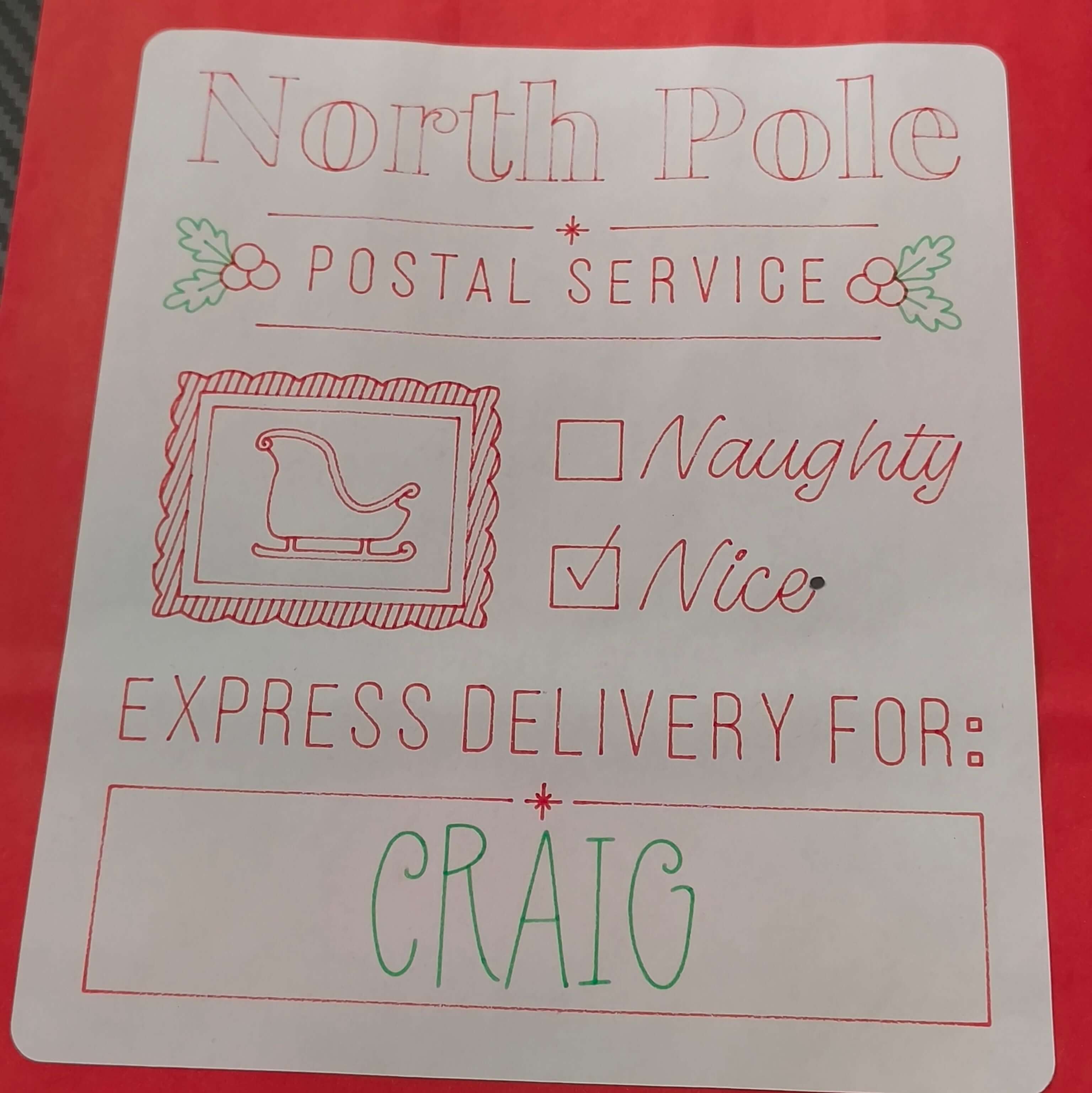 Red Christmas gift bag with a white label reading 'North Pole Postal Service'. The label includes holly decorations, a sleigh icon, checkboxes for 'Naughty' and 'Nice' with 'Nice' checked, and a message that says 'Express Delivery For: CRAIG' in festive red and green lettering.