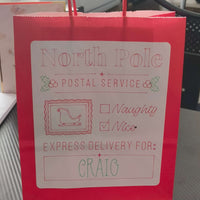 Red gift bag with a large white label that reads 'North Pole Postal Service'. The label features a sleigh icon, holly decorations, checkboxes for 'Naughty' and 'Nice' with 'Nice' checked, and a message that says 'Express Delivery For: CRAIG' in red and green festive lettering.