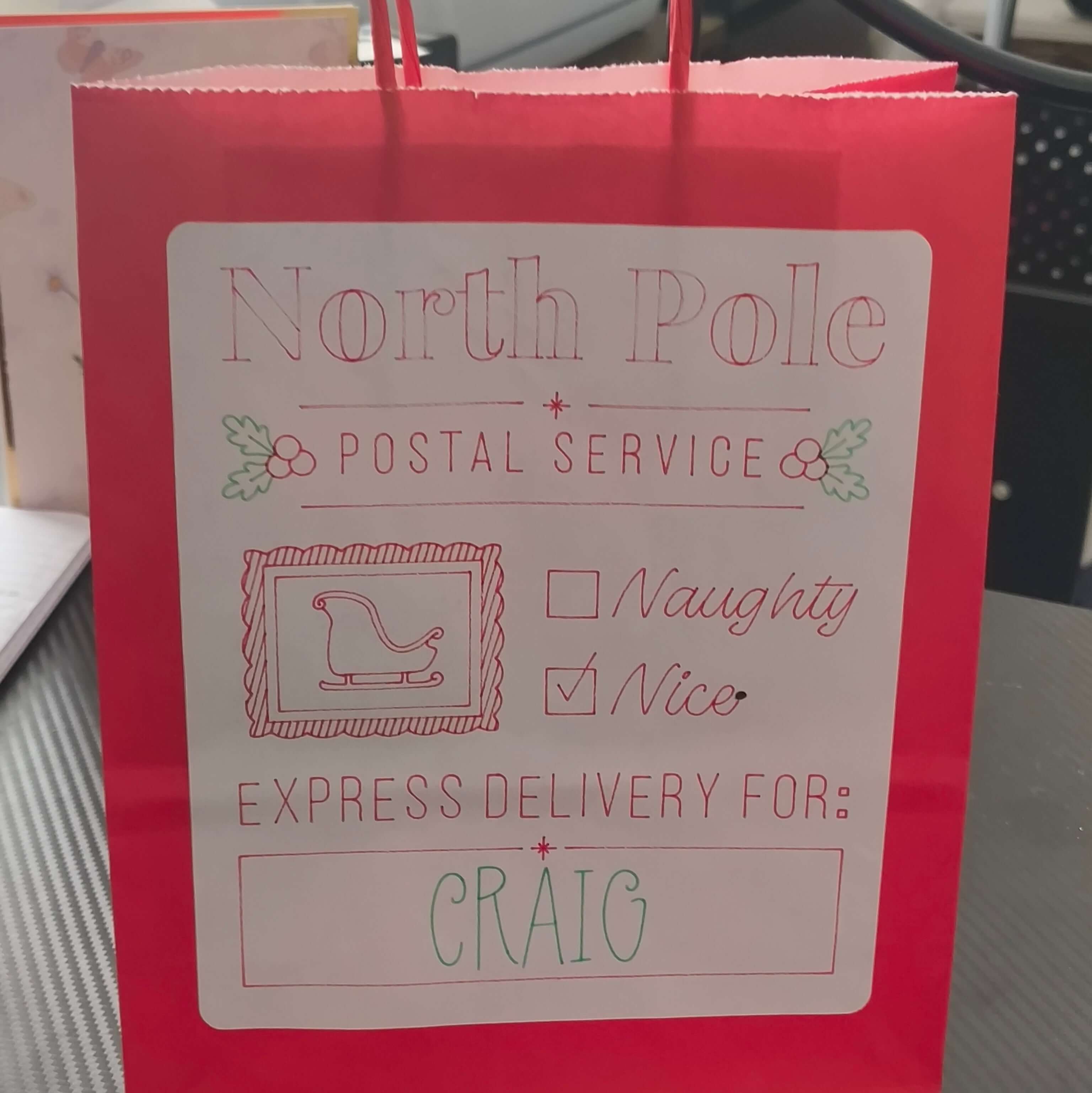 Red gift bag with a large white label that reads 'North Pole Postal Service'. The label features a sleigh icon, holly decorations, checkboxes for 'Naughty' and 'Nice' with 'Nice' checked, and a message that says 'Express Delivery For: CRAIG' in red and green festive lettering.