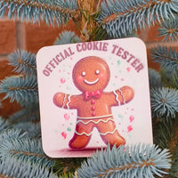 Close-up of festive “Official Cookie Tester” gingerbread coaster with red bow tie design on a spruce tree.