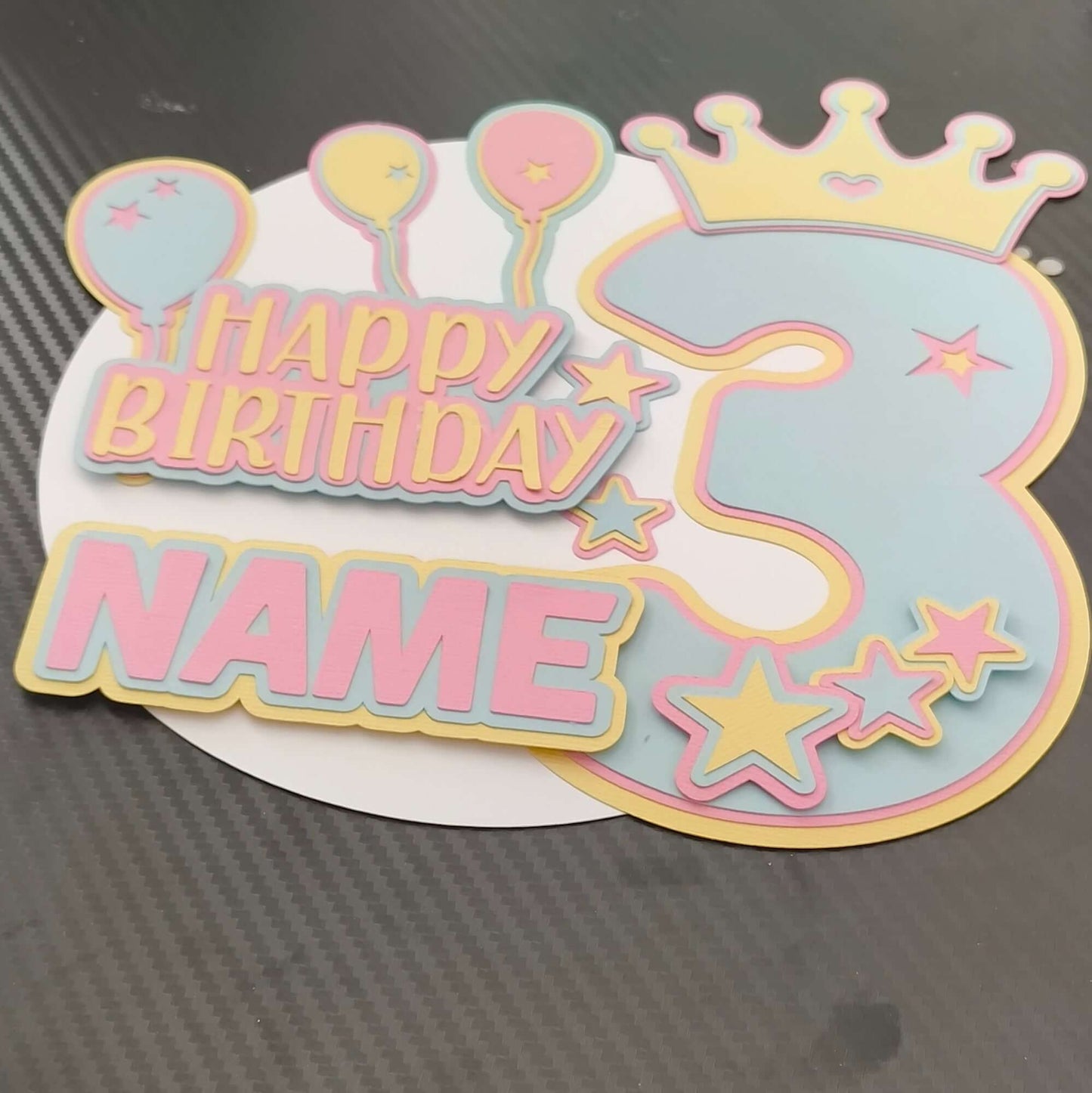 Personalised 3rd birthday cake topper in pastel colours with balloons, crown, and stars, viewed from a slightly angled top-down perspective on a black textured background.