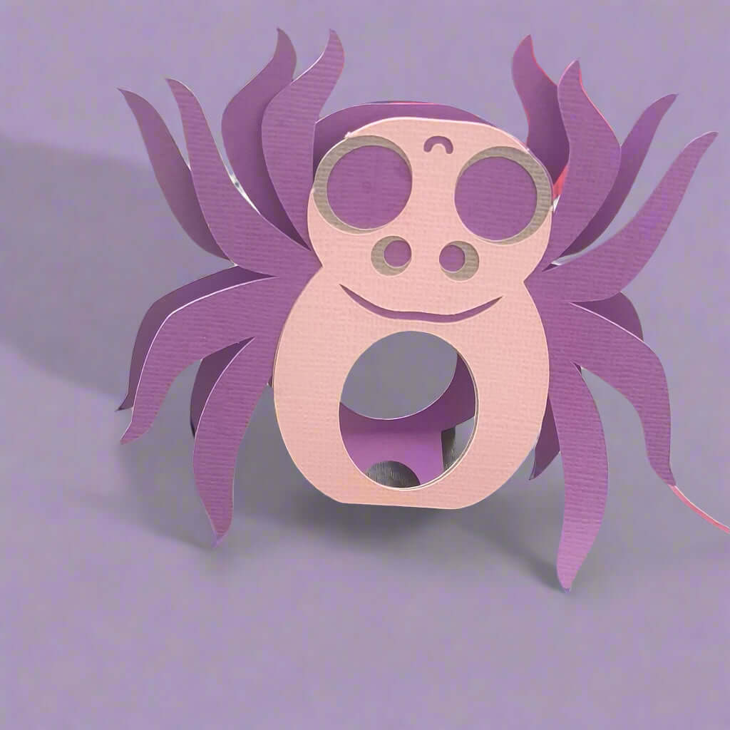 Pastel purple and pink spider-shaped lollipop holder made from textured cardstock, placed on a soft lilac background, perfect for Halloween party favours or handmade spooky gifts.