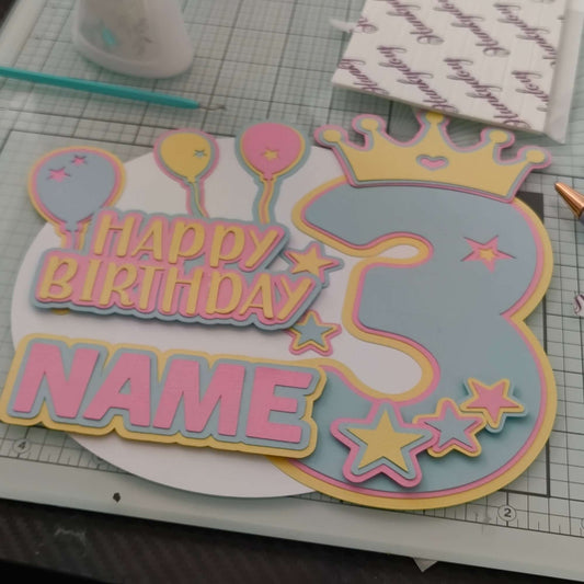 Personalised pastel 3rd birthday cake topper with balloons, crown, stars, and “Happy Birthday” message, shown at a slight angle on a craft mat background.

