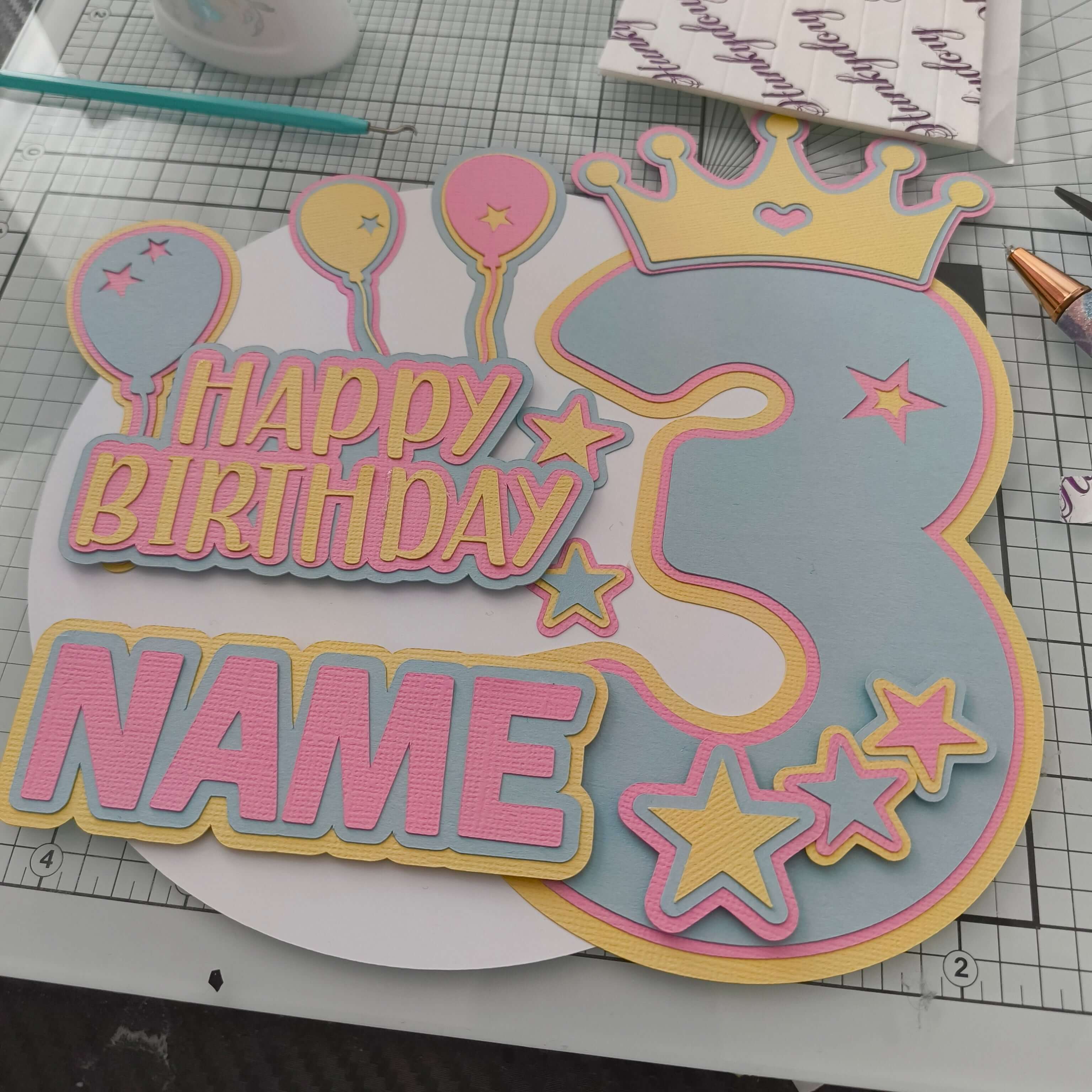 Personalised pastel cake topper for a 3rd birthday, shown from a top-down angle on a craft mat, featuring balloons, stars, crown, and “Happy Birthday” with space for a name.
