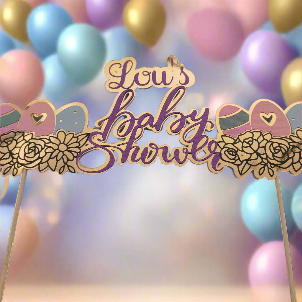 Gold and purple baby shower cake topper reading 'Lou's Baby Shower', decorated with floral and baby-themed elements. The topper is set against a soft-focus background of pastel balloons in blue, pink, lavender, and gold, creating a festive and dreamy atmosphere