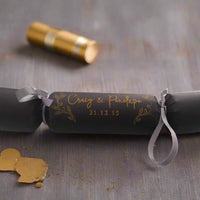 Close-up of black cracker with gold foil text “Craig & Penelope” and date, decorated with silver ribbon and elegant leaf motifs.