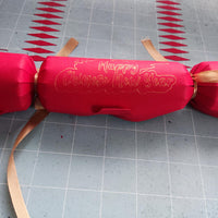 Red cracker with gold foil “Happy Chinese New Year” message, tied with gold ribbon, placed on a craft mat background alongside paper cut-outs.