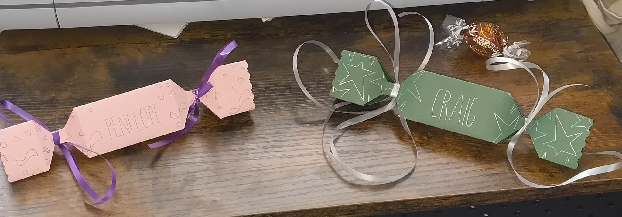 Pair of personalised crackers, one pink with “Penelope” and one green with “Craig,” styled with ribbons and a chocolate truffle for scale.