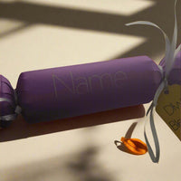 Personalised purple Diwali cracker with white ribbon and 'Diwali Blessings' tag, featuring a space for a name and a small orange diya decoration on a sunlit background.