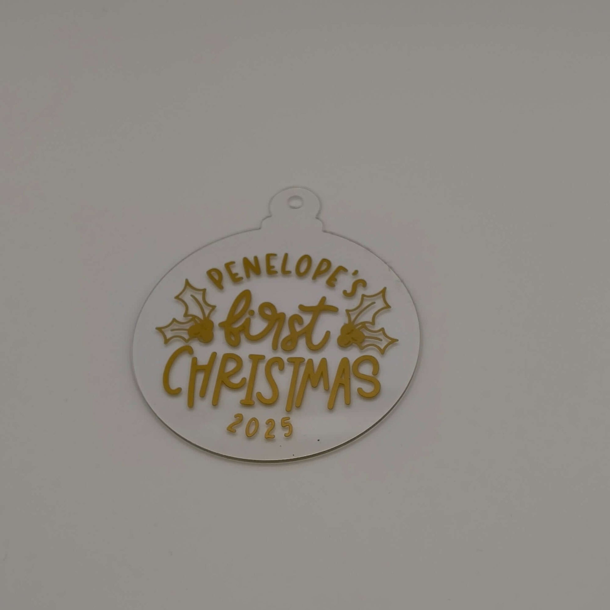 Clear acrylic bauble with gold lettering reading "Penelope’s First Christmas 2025" on plain background.