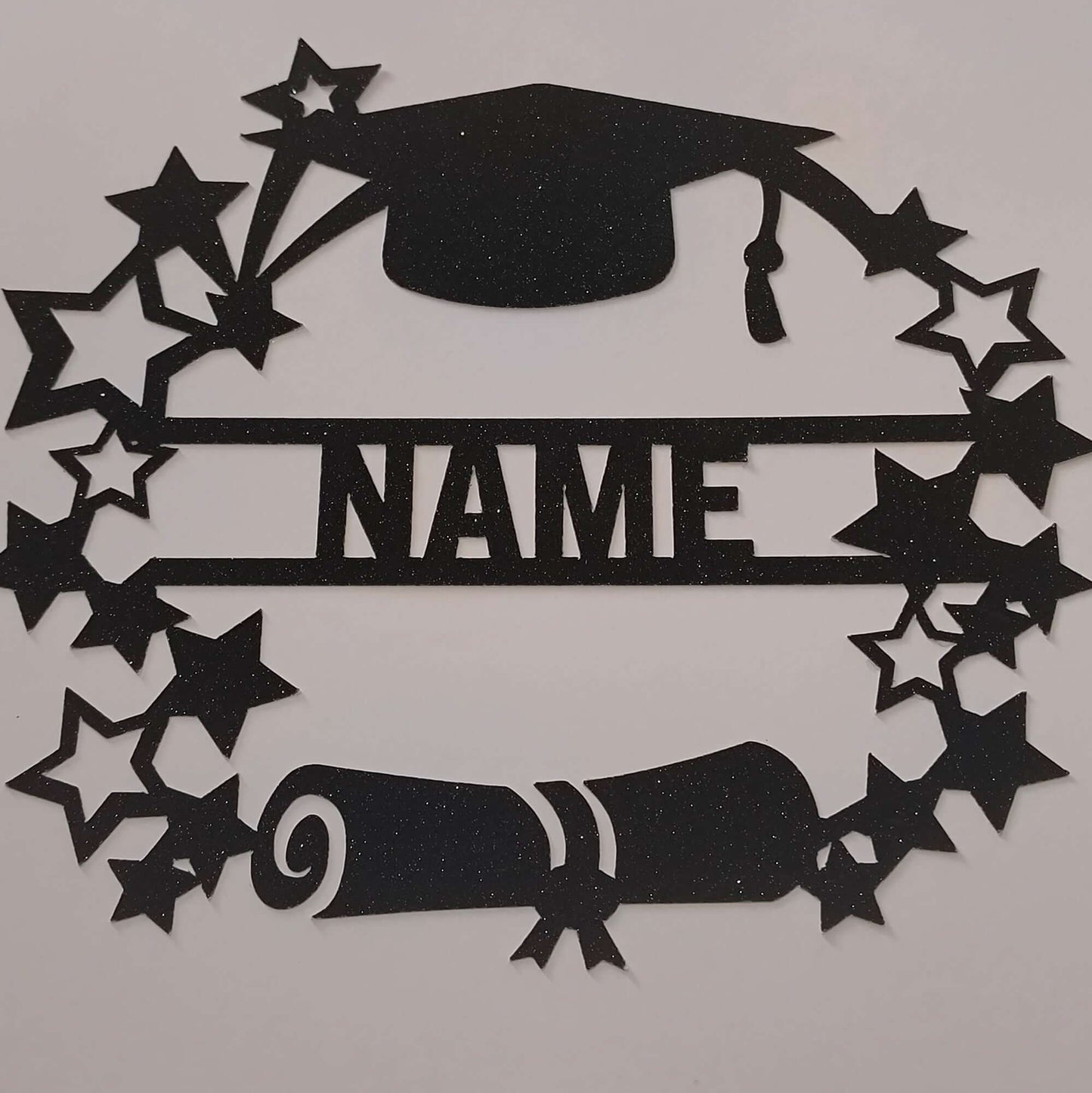 Top-down view of a handmade personalised graduation card topper featuring a black glitter mortarboard, diploma scroll, and star wreath on a white background.