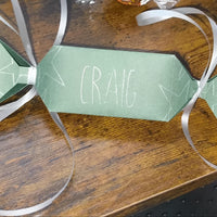 Handmade green personalised cracker with “Craig” in white lettering, star doodle details, and silver ribbon ties, placed on wooden tabletop.