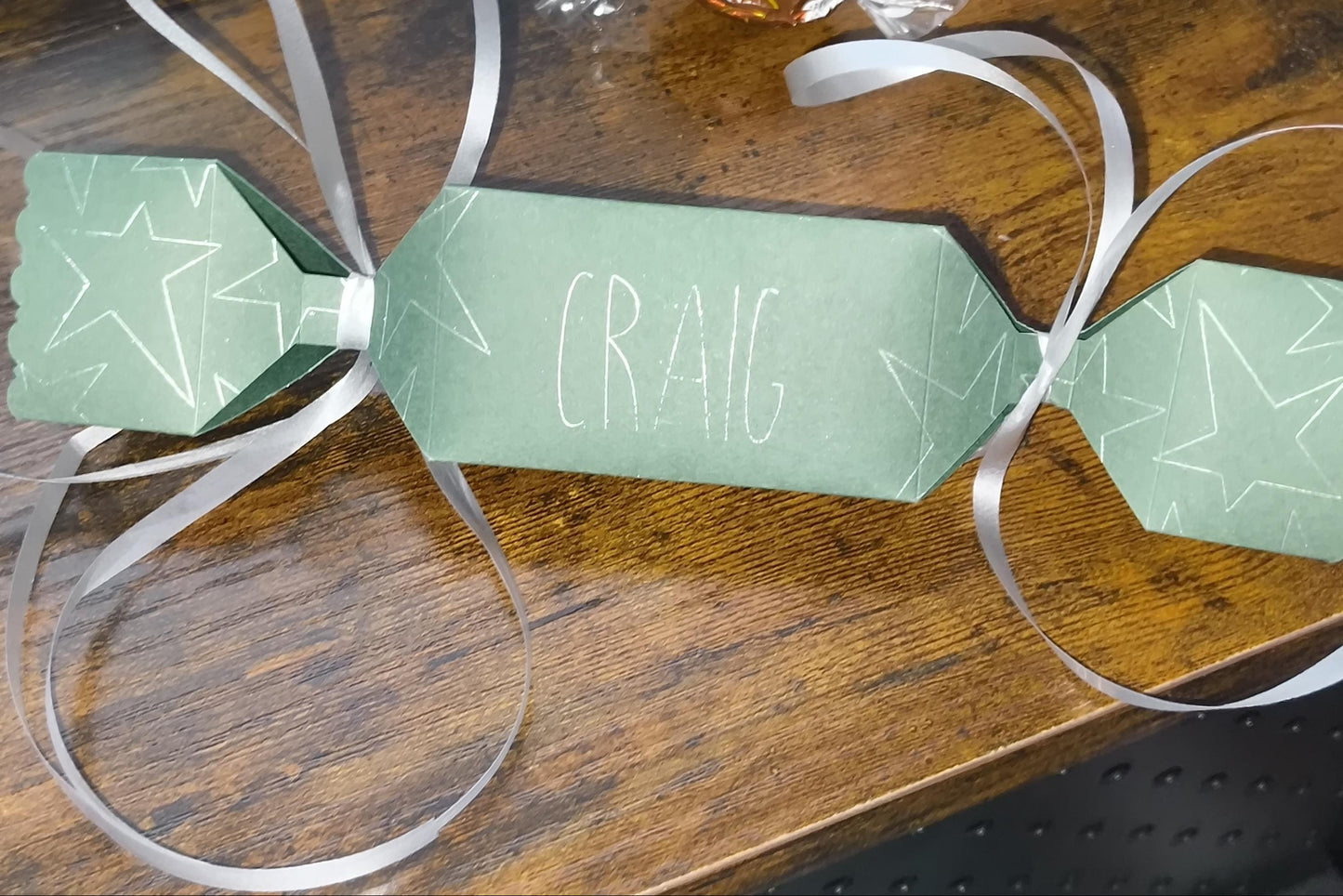 Handmade green personalised cracker with “Craig” in white lettering, star doodle details, and silver ribbon ties, placed on wooden tabletop.