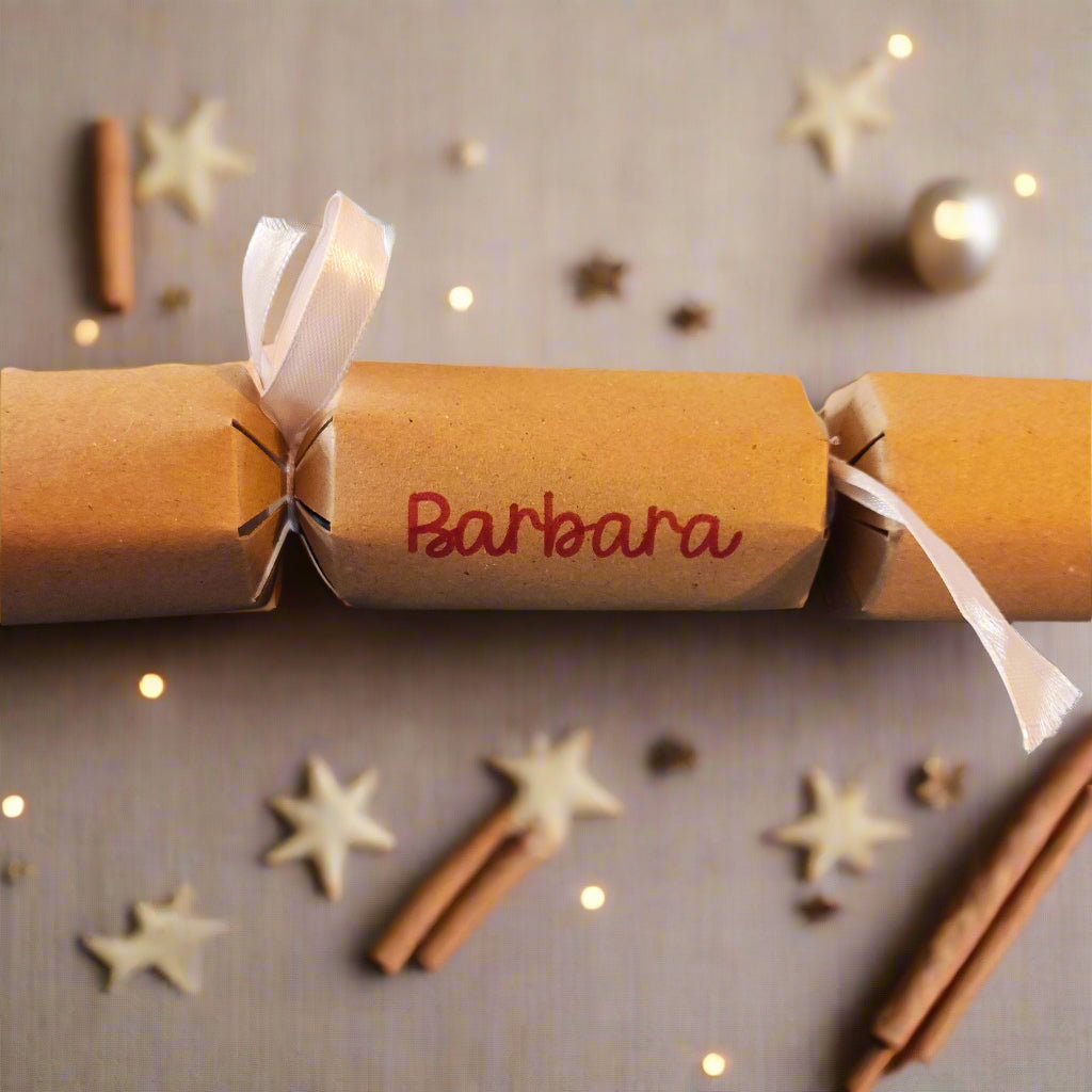 Brown kraft Christmas cracker with white satin ribbon and “Barbara” name in red, styled with cinnamon sticks, star confetti, and warm festive lights.