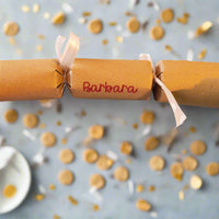 Brown kraft Christmas cracker personalised with “Barbara” in red lettering, tied with white satin ribbon, styled on a table with golden confetti.