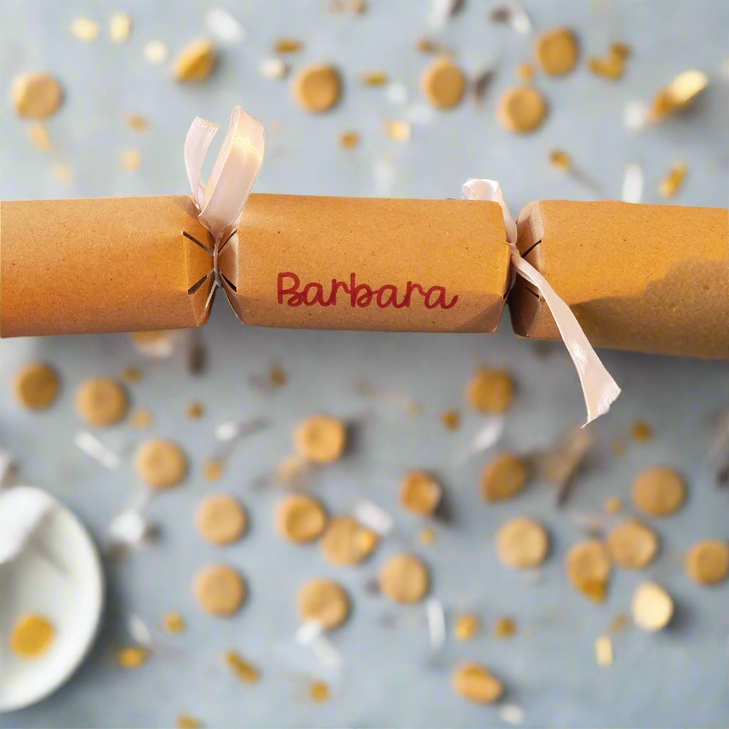 Brown kraft Christmas cracker personalised with “Barbara” in red lettering, tied with white satin ribbon, styled on a table with golden confetti.