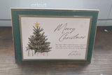 Handmade luxury Christmas card standing upright on a wooden surface. The card features a traditional green textured border with gold foil accents, a central illustration of a Christmas tree topped with a gold star, surrounded by presents. The text reads “Merry Christmas” in elegant script, followed by a heartfelt message and signed “Love the Baxter Family.” 