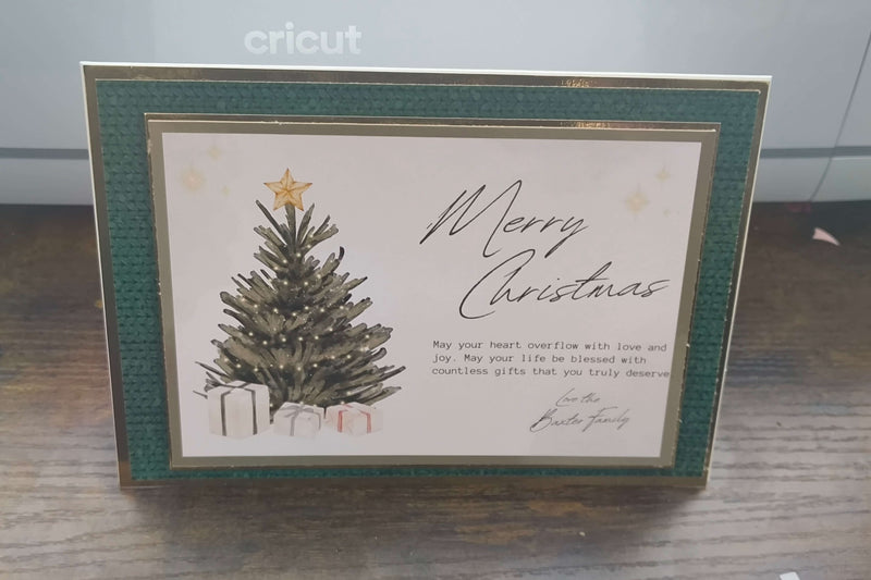 Handmade luxury Christmas card standing upright on a wooden surface. The card features a traditional green textured border with gold foil accents, a central illustration of a Christmas tree topped with a gold star, surrounded by presents. The text reads “Merry Christmas” in elegant script, followed by a heartfelt message and signed “Love the Baxter Family.” 