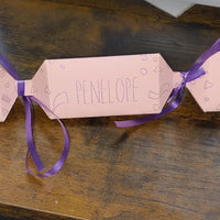 Handmade pink personalised cracker with “Penelope” written across the centre, decorated with geometric doodles and finished with purple ribbon ties.