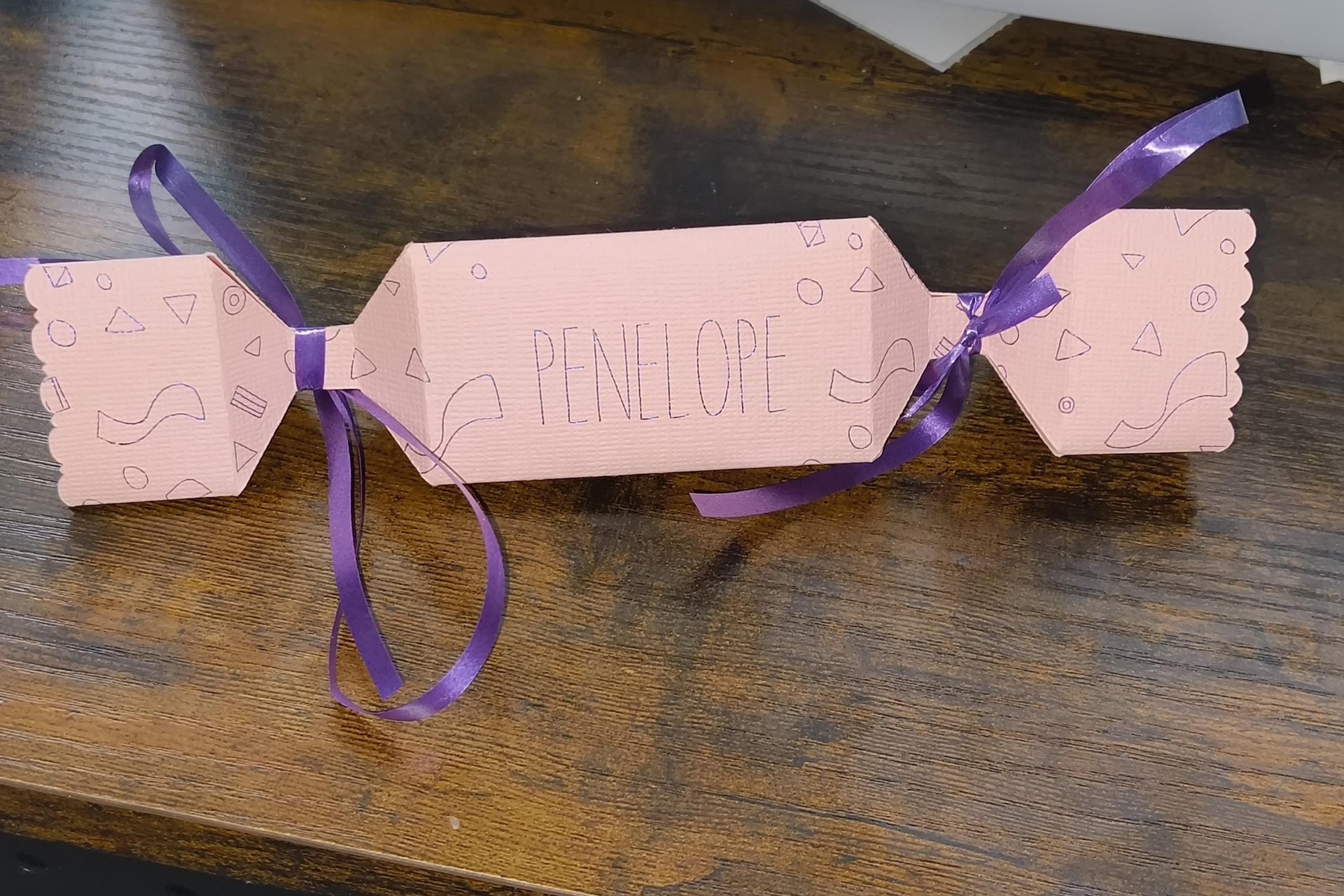 Handmade pink personalised cracker with “Penelope” written across the centre, decorated with geometric doodles and finished with purple ribbon ties.