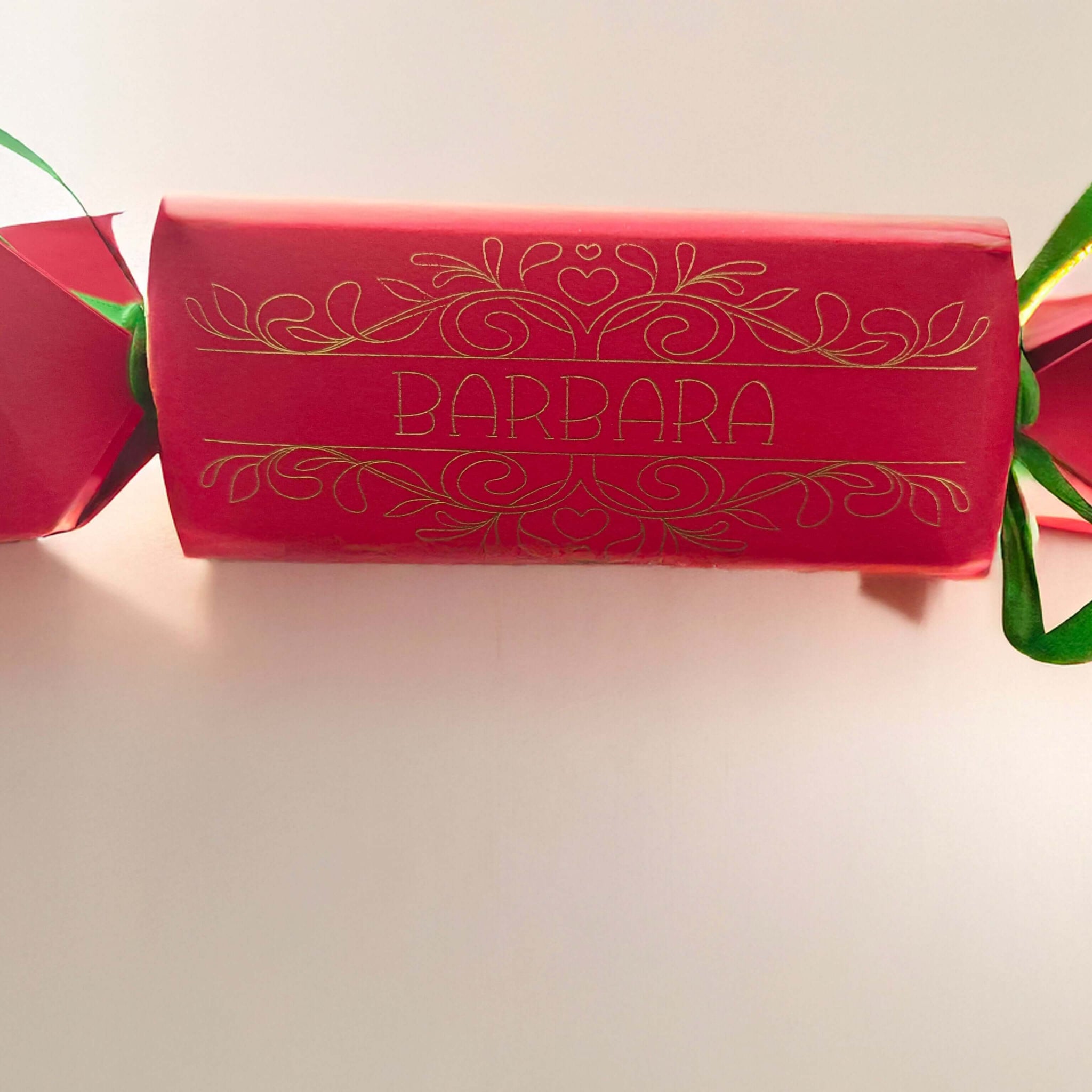 Red cracker with gold foil personalised name “Barbara,” tied with green ribbon, photographed against a pale neutral background with soft light.