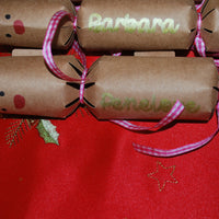 Close-up of personalised kraft reindeer cracker with gold “Penelope” lettering, red nose detail, and pink gingham ribbon, displayed on a red Christmas tablecloth.