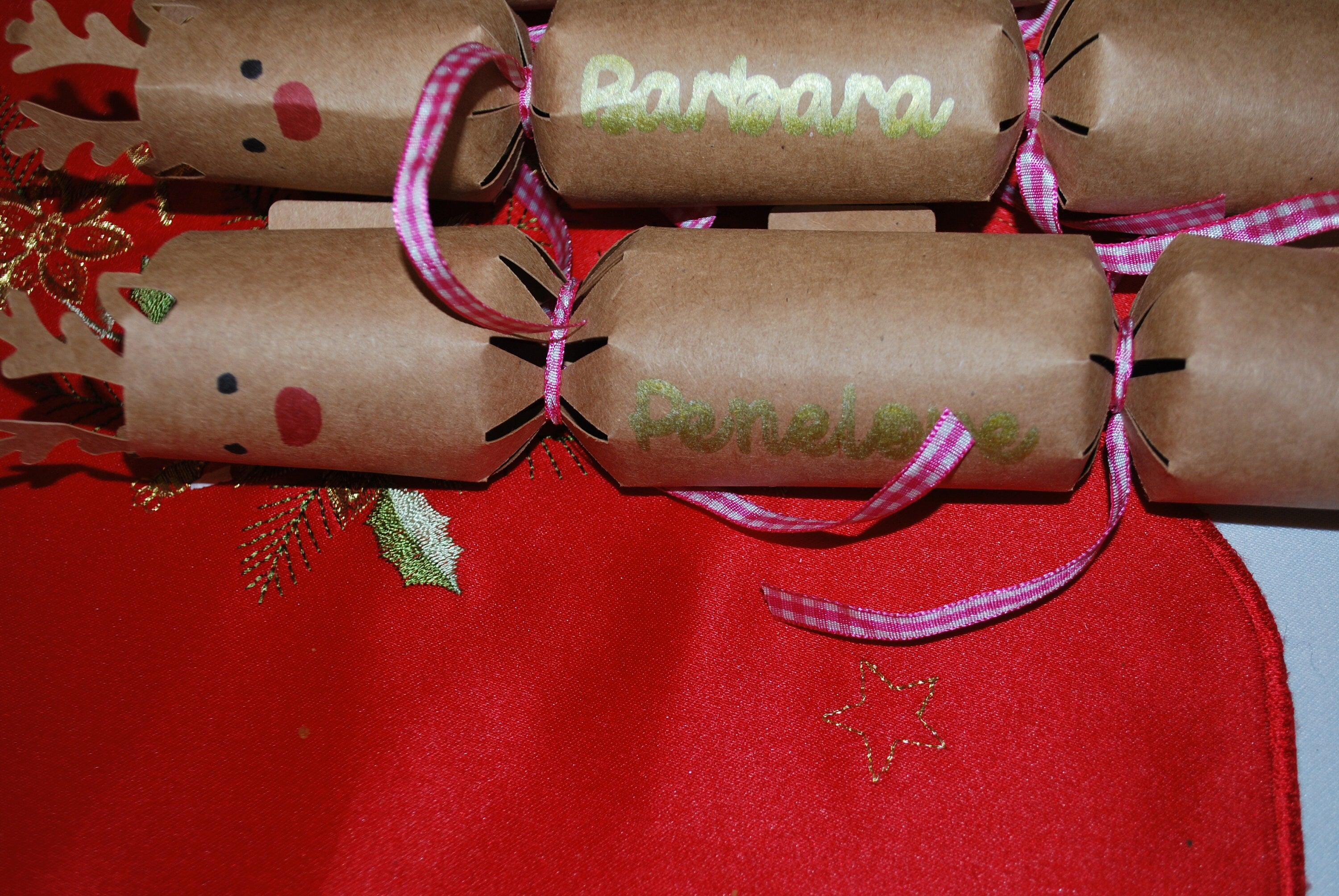 Close-up of personalised kraft reindeer cracker with gold “Penelope” lettering, red nose detail, and pink gingham ribbon, displayed on a red Christmas tablecloth.