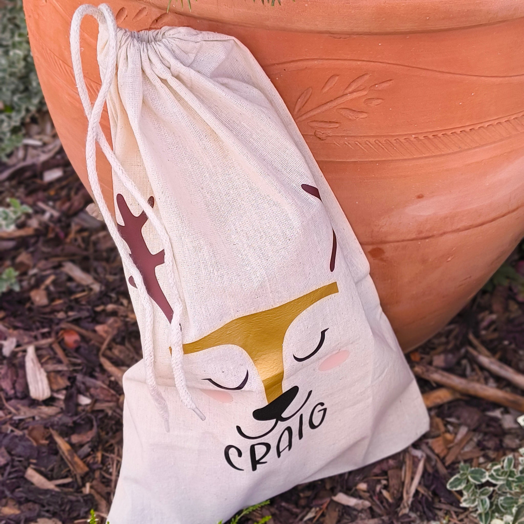Detailed view of personalised reindeer-themed drawstring gift bag with gold and black design, outdoors near plants