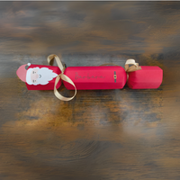 Handmade red Christmas cracker with Santa face, gold ribbon, and personalised name in script text, displayed on rustic wooden background.