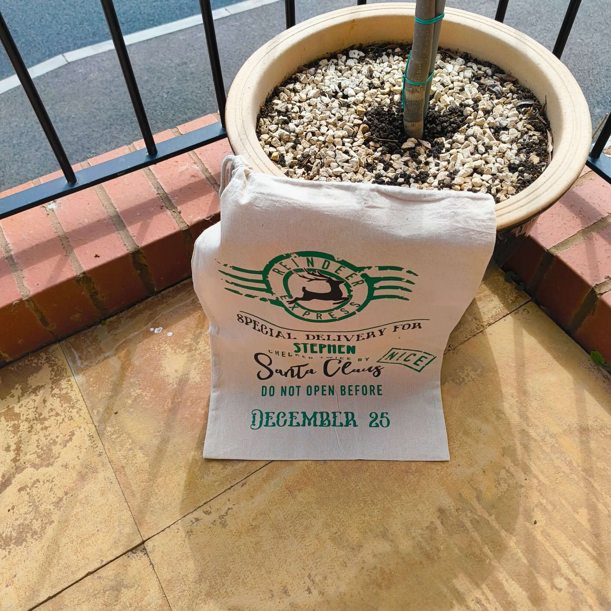 Personalised Christmas Santa sack placed by a plant pot, featuring "Special Delivery from the Reindeer Express" design in green with festive lettering.