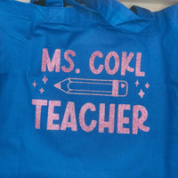 Blue tote bag with pink glitter vinyl text "Ms. Cohl Teacher" featuring a pencil illustration, placed on a wooden surface in a bright home setting.