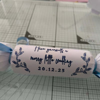 Close-up of white cracker with navy foil wedding message and date, tied with light blue satin ribbon, placed on craft grid surface.