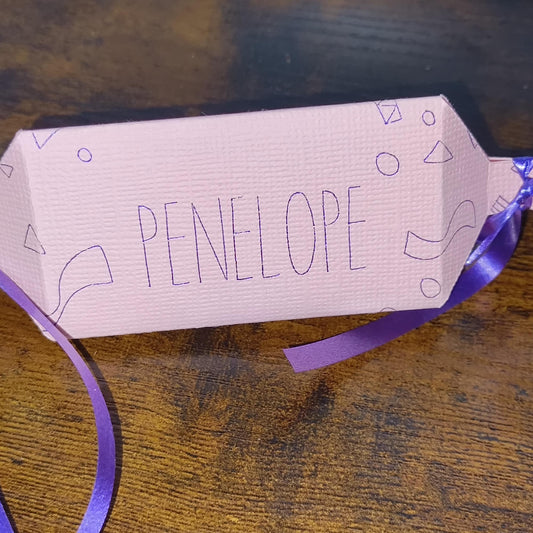 Close-up of pink cracker with “Penelope” in hand-drawn lettering, showing textured cardstock and purple ribbon detail.