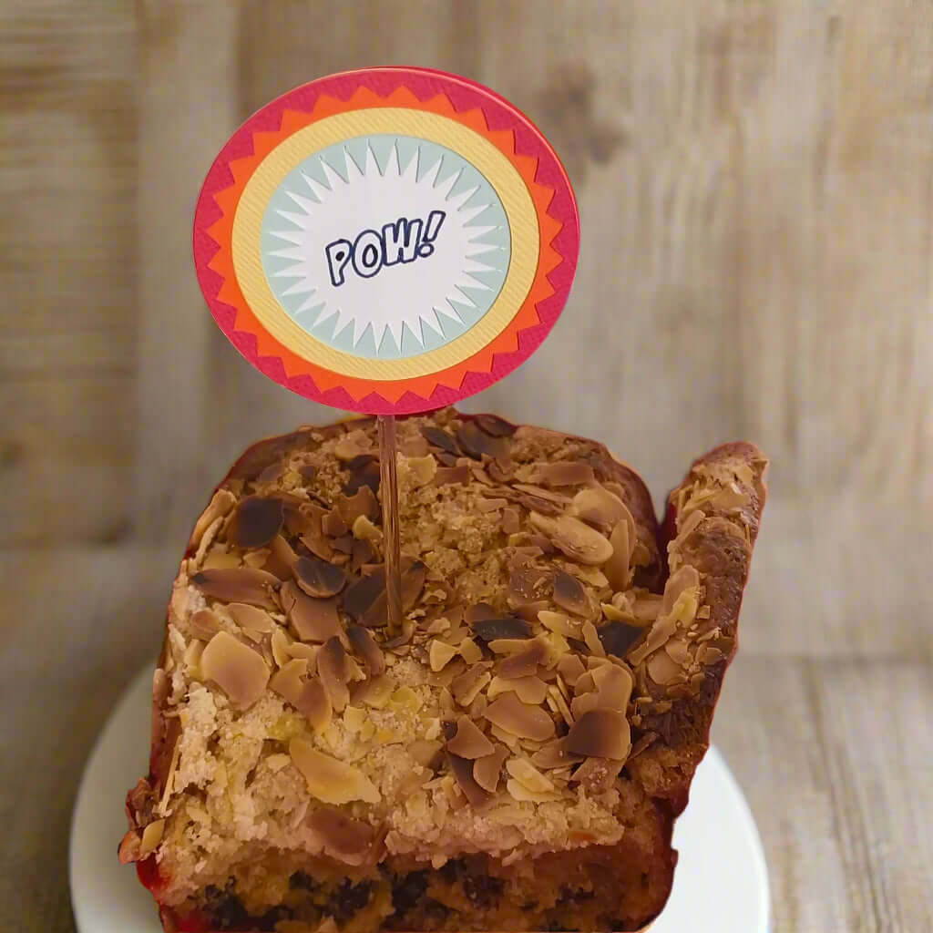 Front-facing view of comic-style 'POW!' cake topper inserted into an almond-topped loaf cake, designed for superhero-themed parties.