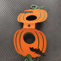 Front close-up of a handmade pumpkin-shaped lollipop holder in bright orange with black bat detail and green stem, photographed from above on a black carbon-textured background.