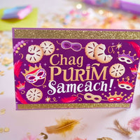 hag Purim Sameach card with gold glitter borders, purple background, festive mask and hamantaschen design, photographed from front angle in a colourful confetti flat lay with close-up detail