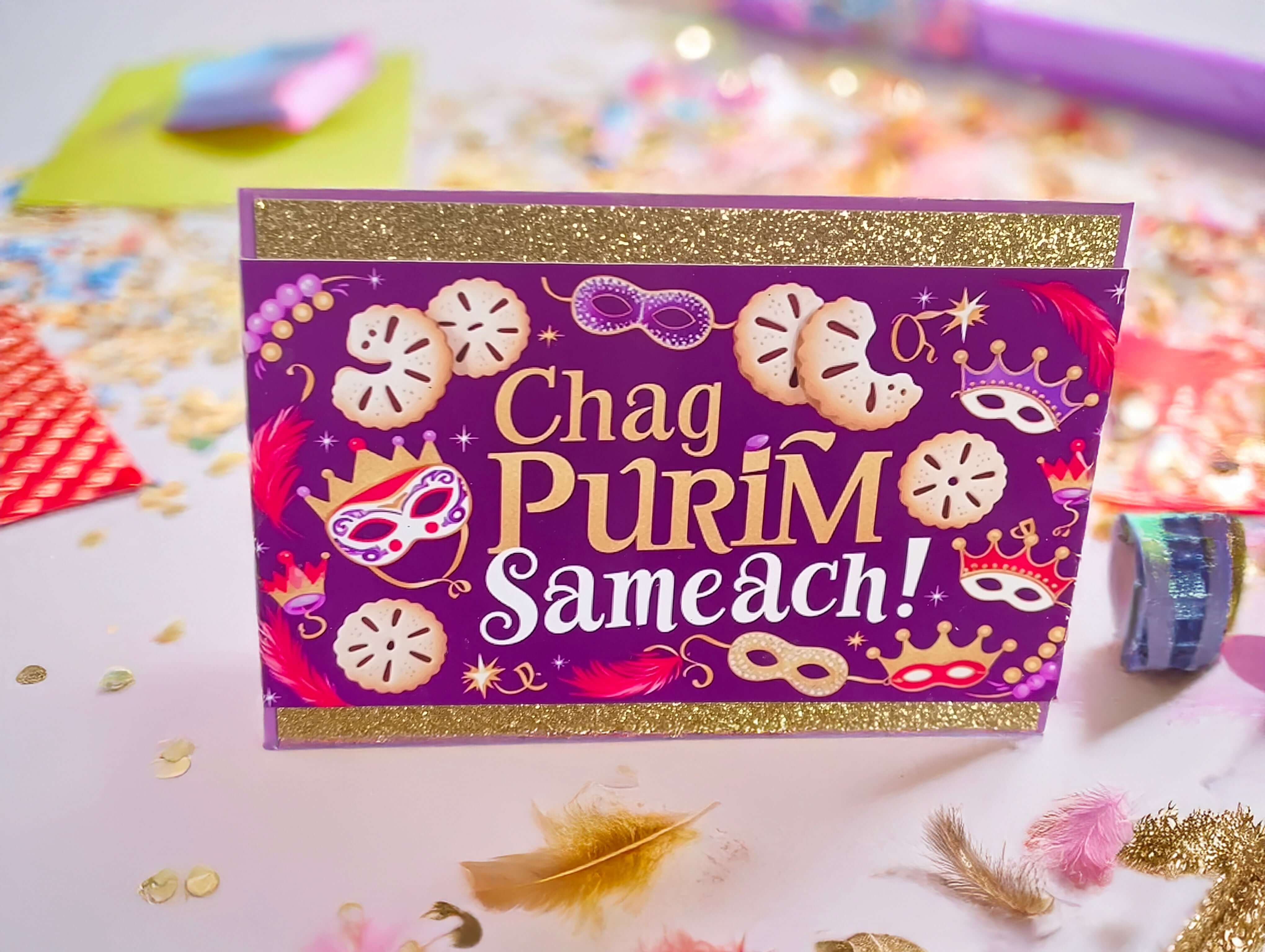 hag Purim Sameach card with gold glitter borders, purple background, festive mask and hamantaschen design, photographed from front angle in a colourful confetti flat lay with close-up detail