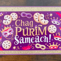 Top-down view of handmade Chag Purim Sameach card with gold glitter trim, purple background, hamantaschen, masks, crowns, and feathers in bold festive colours