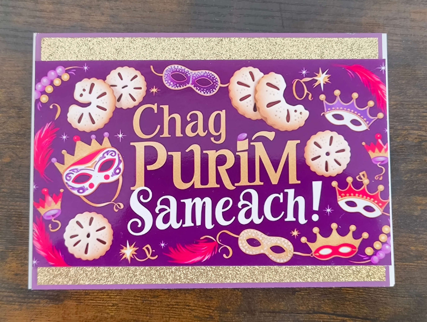 Top-down view of handmade Chag Purim Sameach card with gold glitter trim, purple background, hamantaschen, masks, crowns, and feathers in bold festive colours