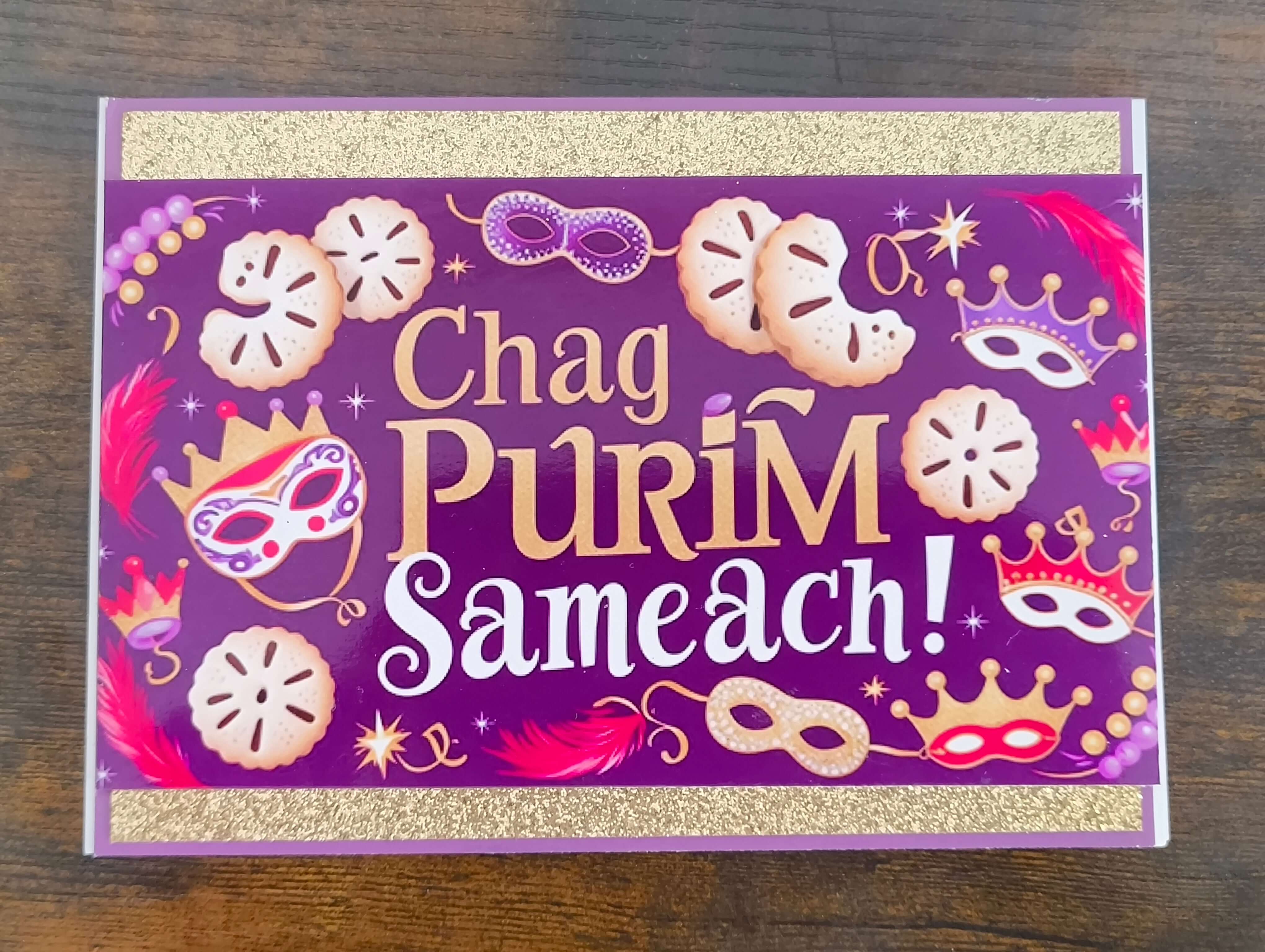 Top-down view of handmade Chag Purim Sameach card with gold glitter trim, purple background, hamantaschen, masks, crowns, and feathers in bold festive colours