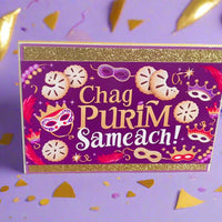 Chag Purim Sameach card with gold glitter borders, purple background, hamantaschen and mask illustrations, front angle close-up on lilac background with gold and confetti details