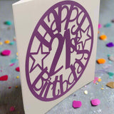 Angled view of a purple papercut 'Happy 21st Birthday' card with stars, displayed on a glittery background scattered with colourful confetti.